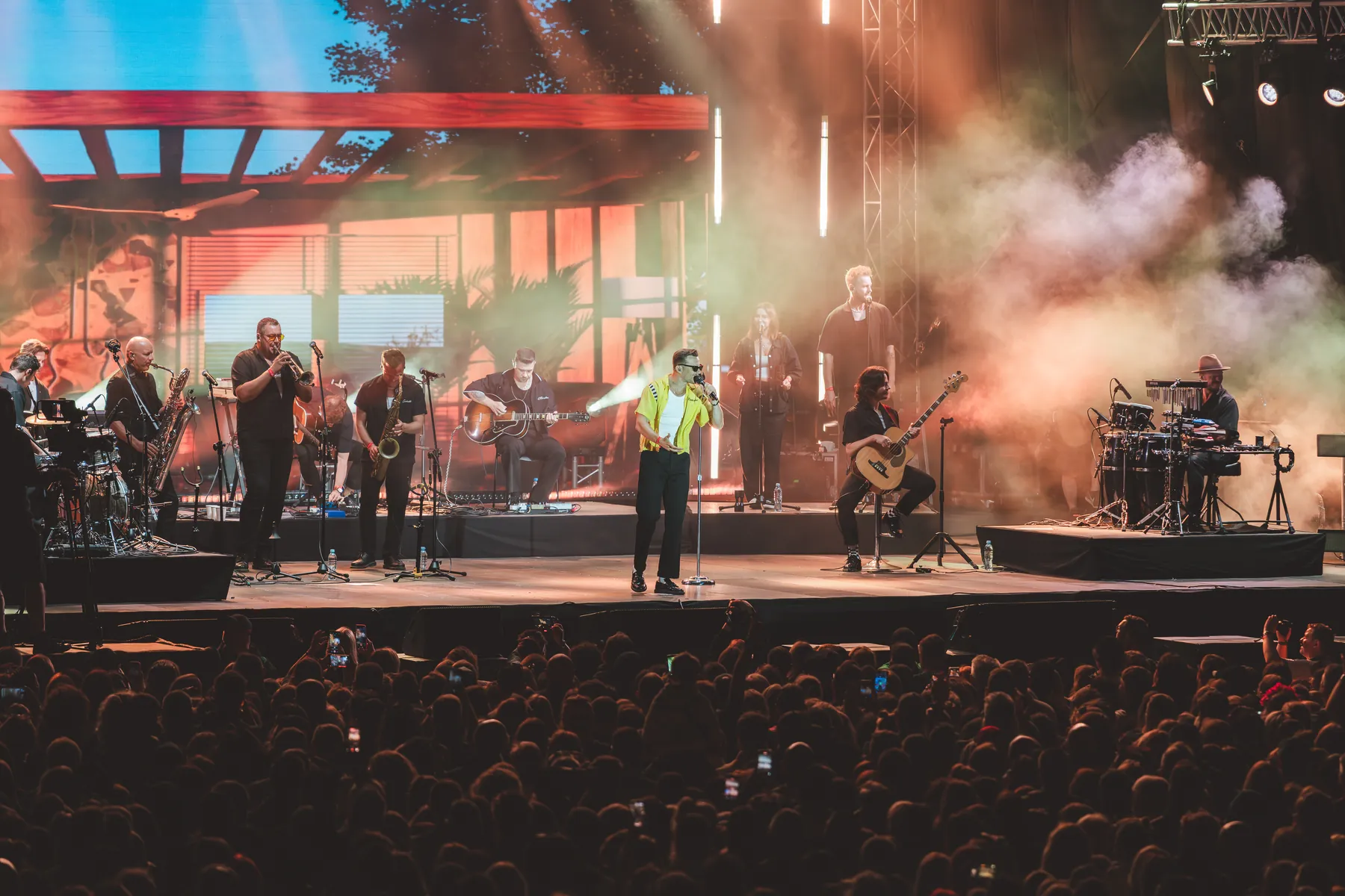 Koncert plenerowy - zespół na scenie w&nbsp;kolorowym dymie i&nbsp;tłum widzów