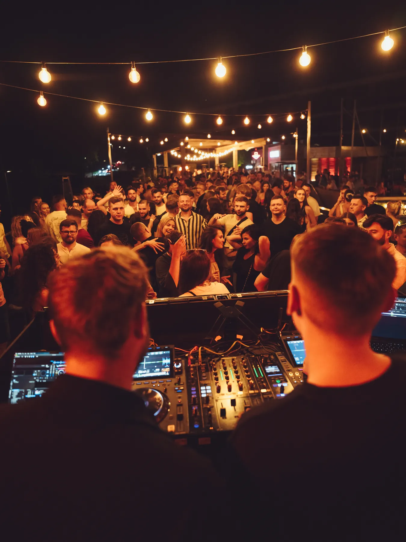 DJ-e za konsoletą przed tłumem pod girlandą żarówek